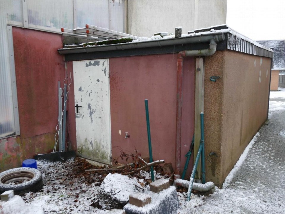 Rückansicht eines Gebäudes mit roter Fassade, weißer Tür und Schneebedeckung; umliegende Gegenstände wie Reifen und Werkzeuge