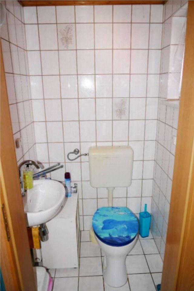 Badezimmer mit weiß gefliesten Wänden, Toilette mit blauem Sitz, Waschbecken und Toilettenbürste