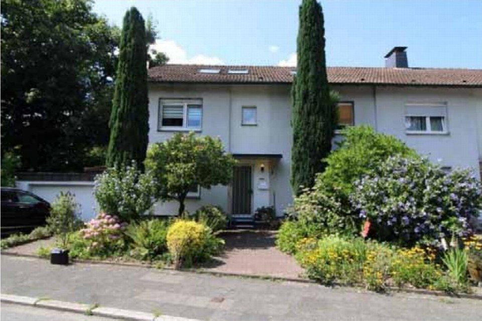 Vorderansicht eines Zweifamilienhauses mit Eingang, Fenstern, Ziegeldach und vorgelagertem Garten mit Sträuchern und Bäumen