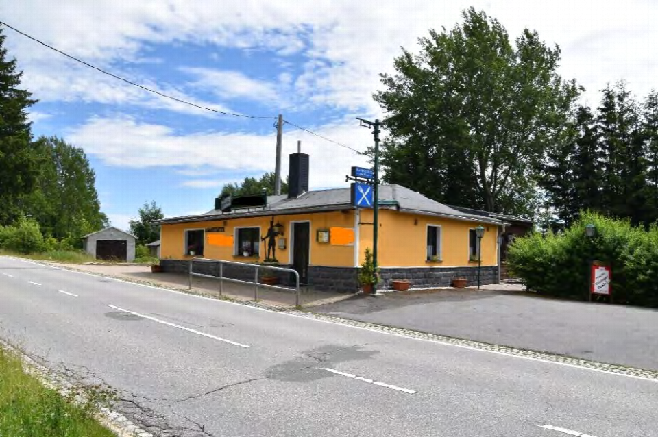 Gelbes Gebäude mit dunklem Dach an der Eibenstocker Straße 91 in Johanngeorgenstadt, Ansicht von vorne mit Straße und Bäumen im Hintergrund