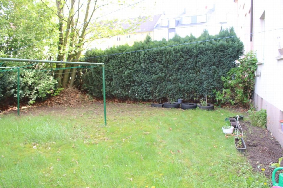 Grüner Gartenbereich mit Rasen, Hecken, Bäumen und Gartenutensilien wie Reifen und Pflanzkübeln an der Seite eines Gebäudes