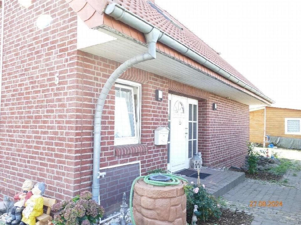 Eingangsbereich eines Backsteinhauses mit weißer Tür, Fenster und Regenrohr; im Vordergrund Dekoration und Gartenelemente