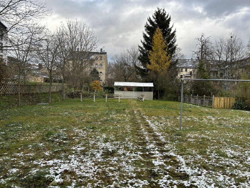 Grüner Gartenbereich mit vereinzeltem Schnee, kahlen Bäumen, Holzzaun und kleinem Schutzdach im Hintergrund