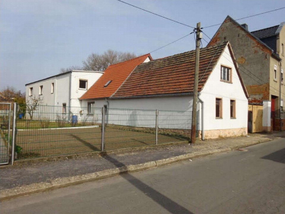 Seitenansicht eines weißen Gebäudes mit rotem Ziegeldach in Teuchern OT Nessa, Kapellenende 69, hinter einem Metallzaun und neben einer Straße