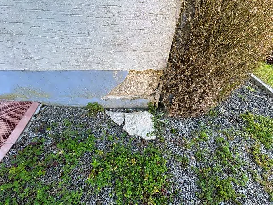 Gartenbereich mit Kies, Unkraut und beschädigter Betonsockel neben einer Hauswand in Altena, Alter Weg 36
