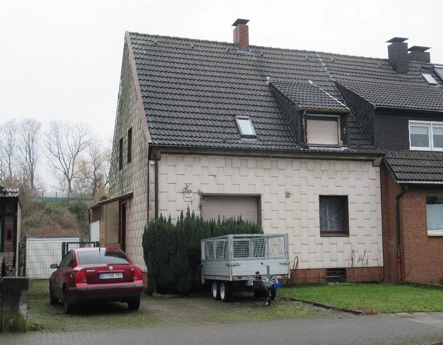 Seitenansicht eines Einfamilienhauses mit Satteldach, Garagenstellplatz und geparktem Auto sowie Anhänger im Vorgarten