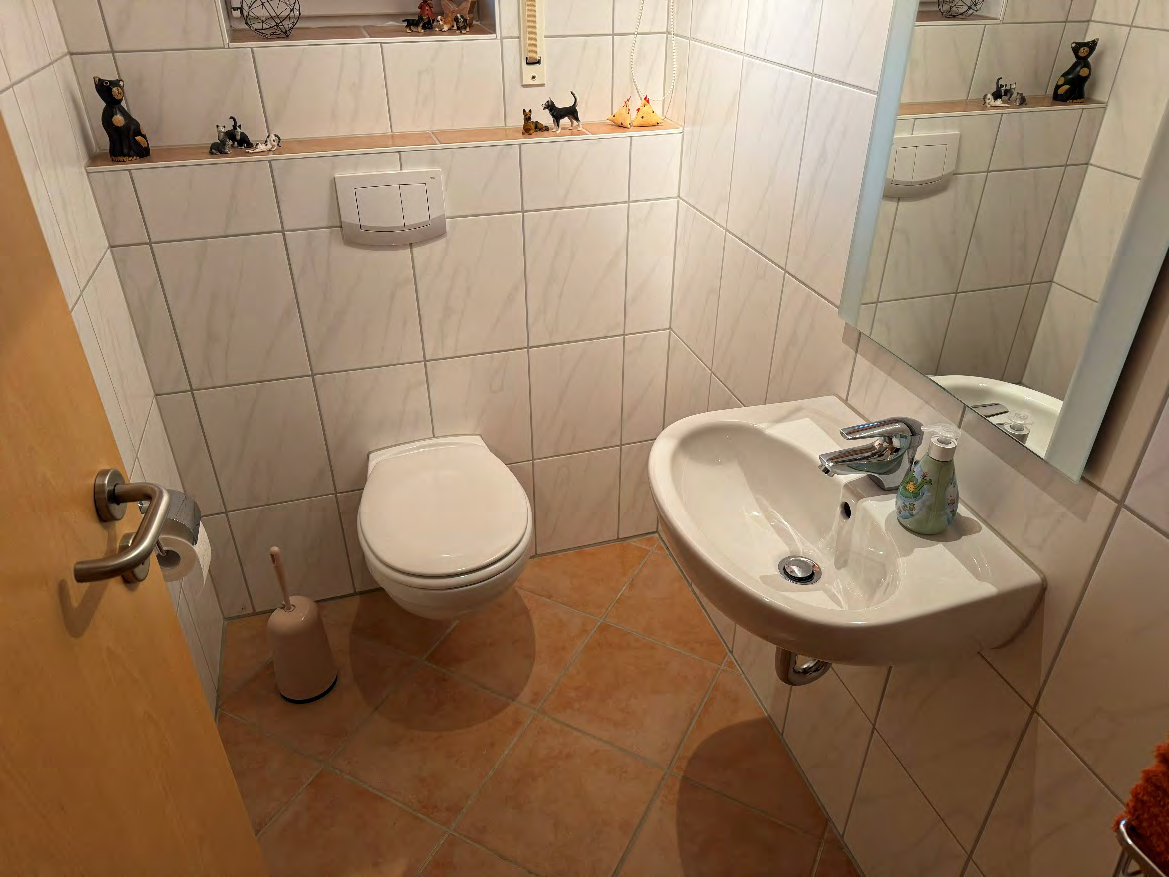 Badezimmer mit weiß gefliesten Wänden, Wandwaschbecken, WC, Spiegel und braunen Fliesenboden. Dekoobjekte auf der Ablage über dem WC
