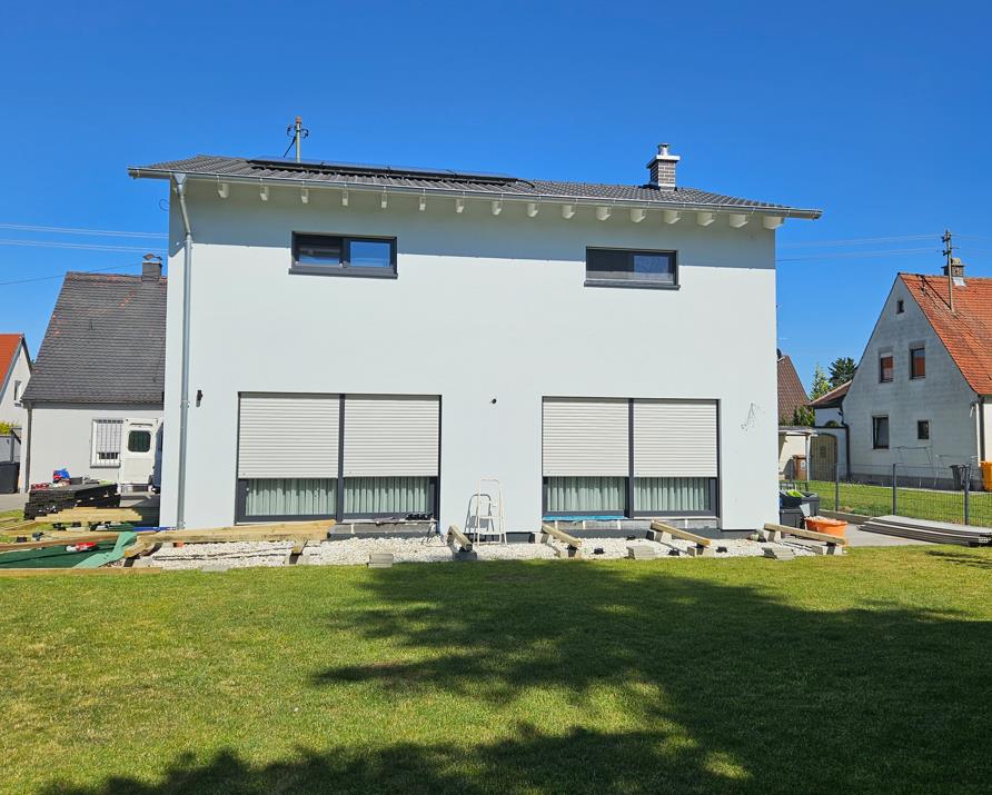 Einfamilienhaus in Augsburg, Falkenweg 7a, mit weißer Fassade, dunklem Dach und Rollläden, vorne Grünfläche, im Hintergrund weitere Häuser