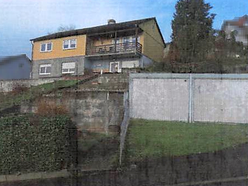 Seitenansicht eines Einfamilienhauses in Wiesenbach, Bergstraße 9, mit gelb-grauer Fassade, Balkon und Betonmauer im Vordergrund