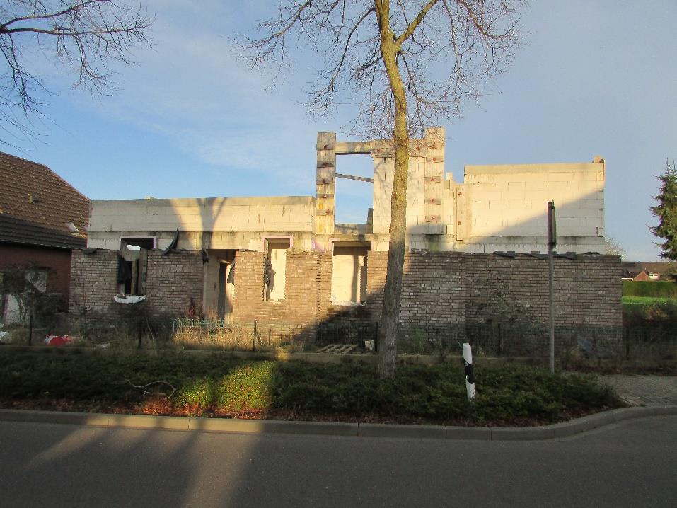 Vorderansicht eines zweifamilienhauses im Rohbauzustand mit sichtbaren Ziegel- und Betonwänden, teilweise ohne Dach, umgeben von einer Hecke und einem Baum