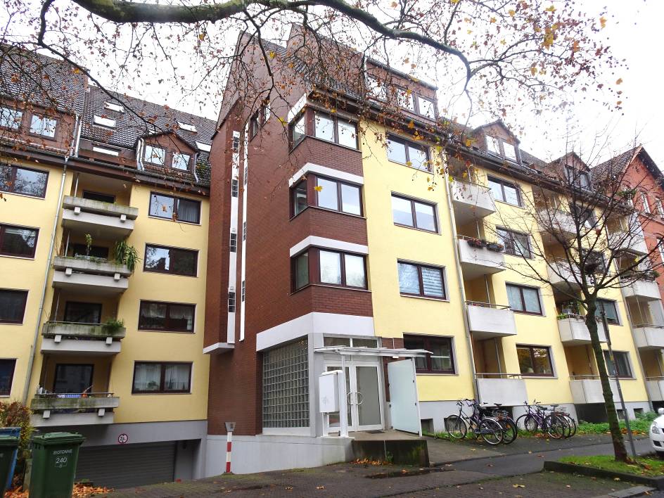 Eingangsbereich eines Mehrfamilienhauses mit weißer Tür, Glasblockwand und Vordach, umgeben von gelb- und braun verputzten Fassadenabschnitten