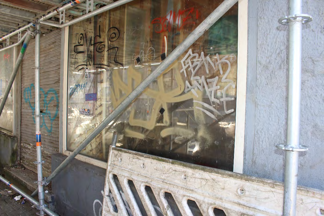 Seitenansicht eines Gebäudes mit Graffiti an der Fensterscheibe, Baugerüst und Holzbarriere im Vordergrund