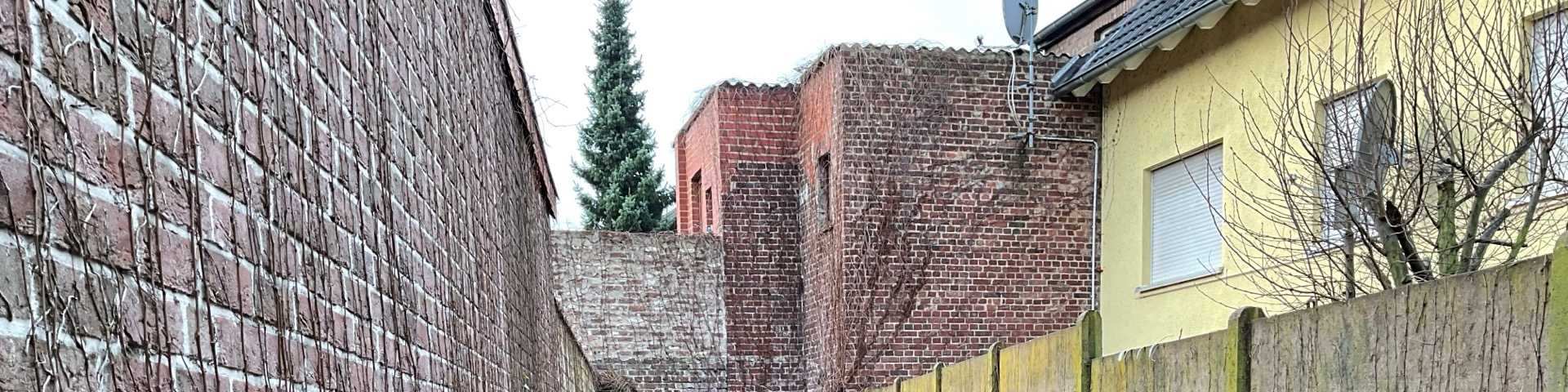 Seitenansicht eines Backsteinhauses mit angrenzenden Mauern, einem Holzzaun und kahlen Bäumen im Vordergrund. Im Hintergrund ist ein Nadelbaum sichtbar