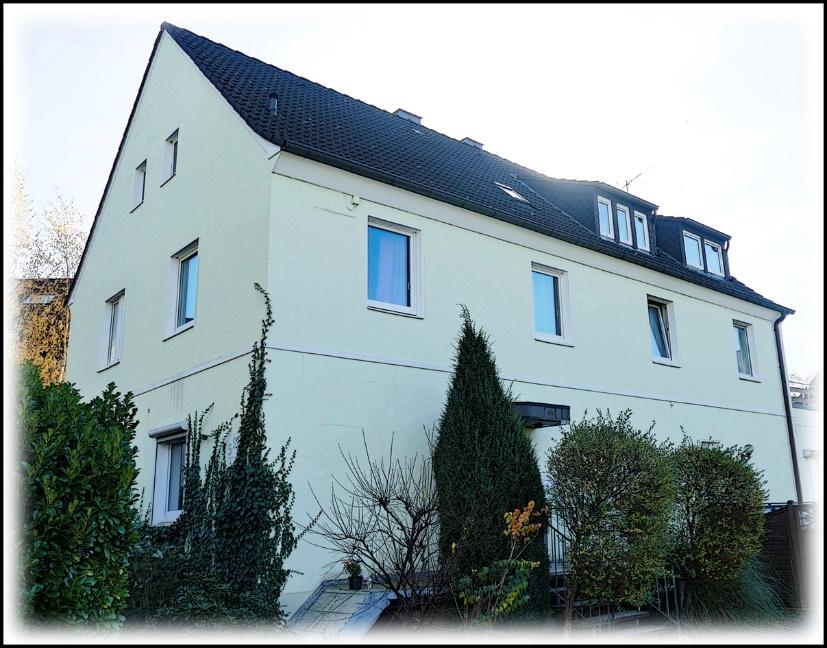 Seitenansicht eines Mehrfamilienhauses mit hellgrüner Fassade, dunklem Dach und Fenstern, umgeben von Sträuchern und Bäumen