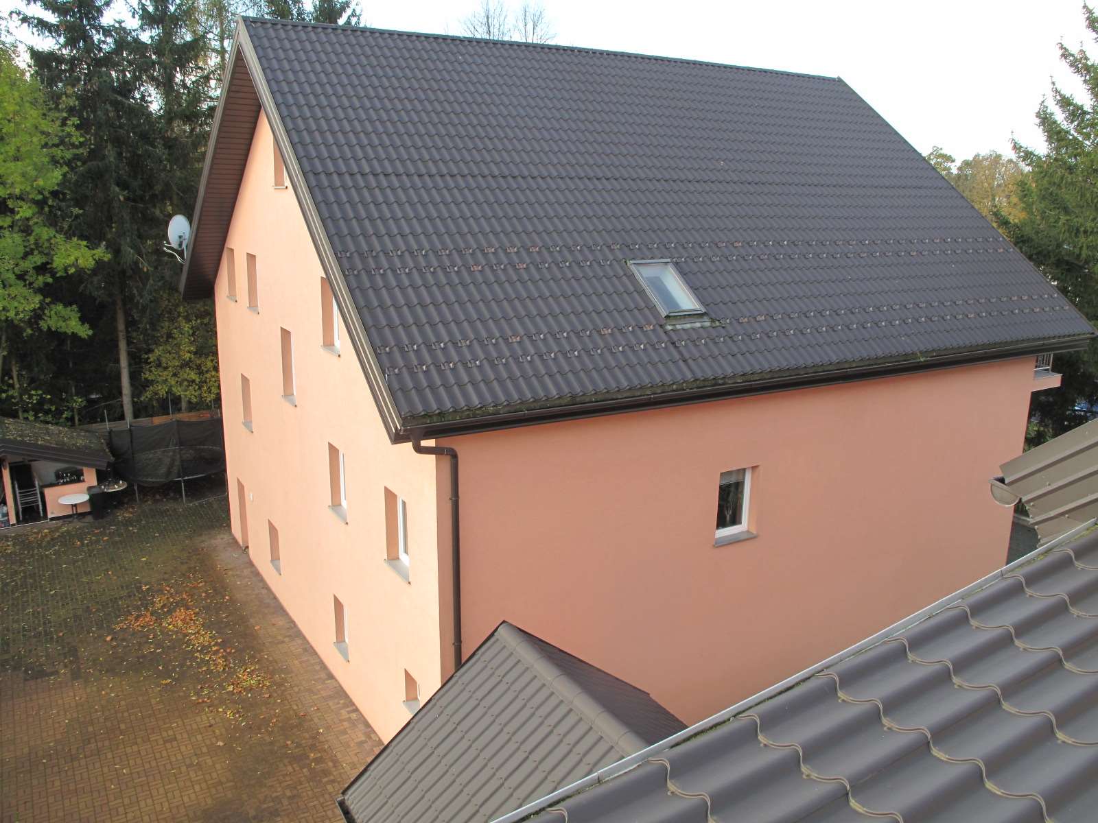 Seitenansicht eines mehrstöckigen Gebäudes mit rosa Fassade und dunklem Wellendach, umgeben von Bäumen und einem gepflasterten Hof