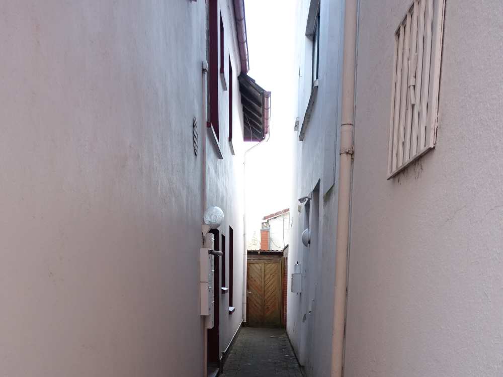 Schmale Gasse zwischen zwei Gebäuden mit sichtbaren Wänden, Fenstern und einem Tor am Ende, Ansicht der Seitenfassade des Wohn-/Geschäftshauses in Edewecht