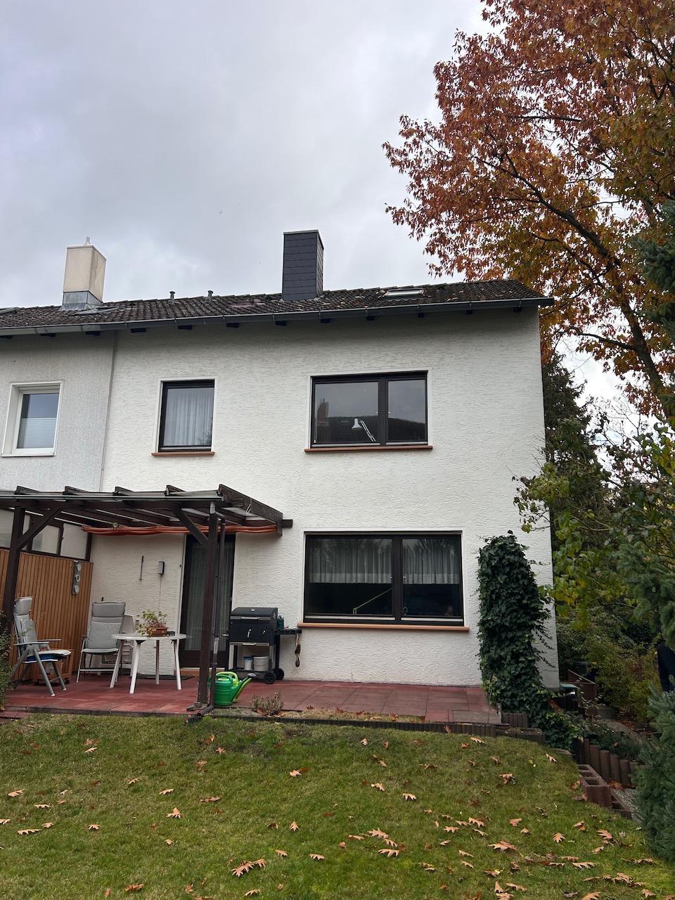 Rückansicht eines zweistöckigen Wohnhauses mit Terrasse, Garten und Gartenmöbeln. Dach mit Schornsteinen, Fenster mit dunklen Rahmen