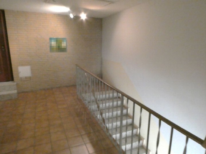 Treppenhaus mit Fliesenboden, Metallgeländer und beigefarbener Wand; im Hintergrund Wand mit Fliesenverkleidung und Bild; Beleuchtung an der Decke