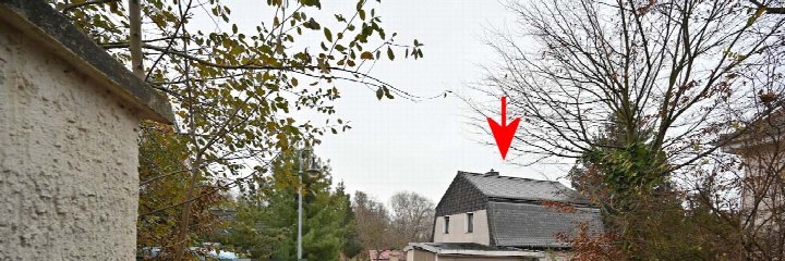 Außenansicht eines Einfamilienhauses von der Seite, mit grauem Dach, weißen Wänden und umgebenden Bäumen; roter Pfeil zeigt auf das Dach