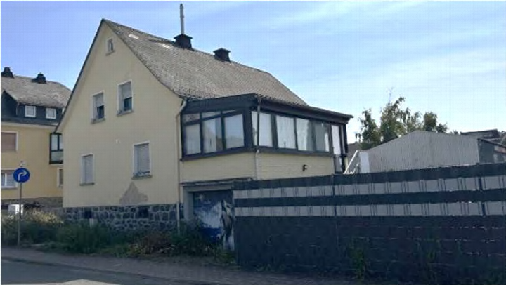 Zweifamilienhaus in Wetzlar-Hermannstein, Wiesenstraße 4, mit gelber Fassade, dunklem Satteldach und Erker. Teilweise verputzte Mauer und Zaun im Vordergrund