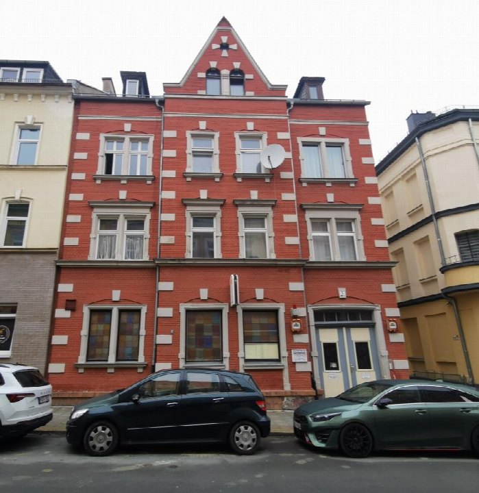 Rotes Backsteingebäude mit weißen Verzierungen, mehreren Fenstern und Eingangstür, Sedanstraße 5, Hof, 95028. Davor geparkte Autos
