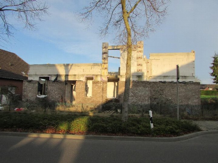 Vorderansicht eines zweifamilienhauses im Rohbauzustand mit sichtbaren Ziegel- und Betonwänden, teilweise ohne Dach, umgeben von einer Hecke und einem Baum