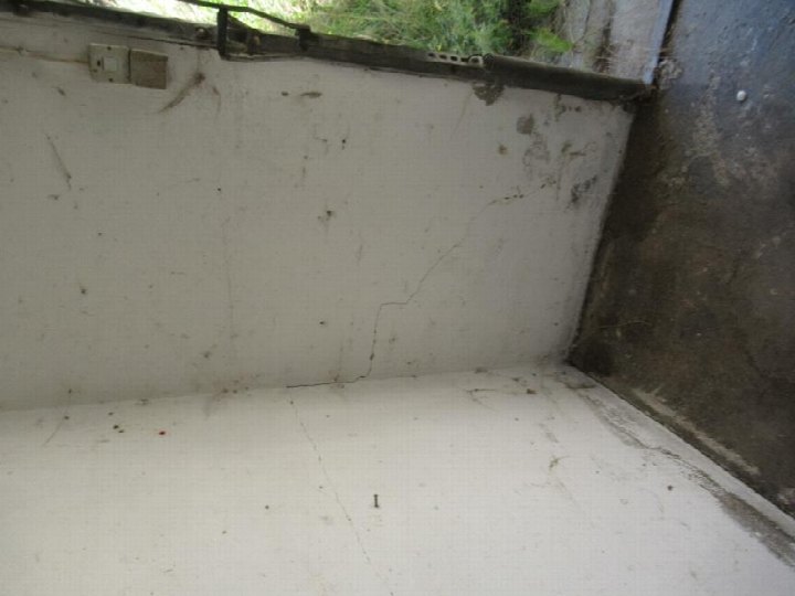 Innenansicht einer Garage mit weiß gestrichener Wand, sichtbaren Rissen und einer elektrischen Steckdose. Teilweise sichtbarer Betonboden und Außenbereich mit Pflanzen