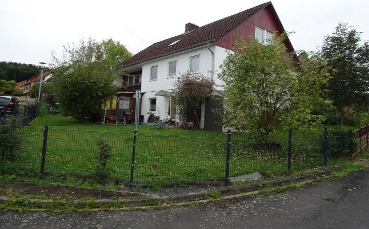Einfamilienhaus mit weißer Fassade und rotem Dach, umgeben von Grünfläche und Metallzaun, an der Ecke Grüner Weg 2 in Hardegsen, 37181