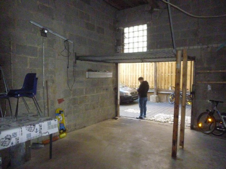 Garage mit Betonboden und Ziegelwänden, geöffnete Tür zum Außenbereich, darin ein Mann, ein Auto, Fahrräder und ein Skateboard an der Wand