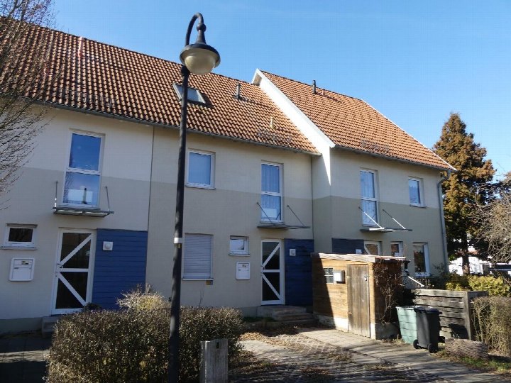Zweistöckiges Reihenmittelhaus mit Satteldach, hellen Fassaden und blauen Türen, vorne mit Straßenlaterne und gepflastertem Weg