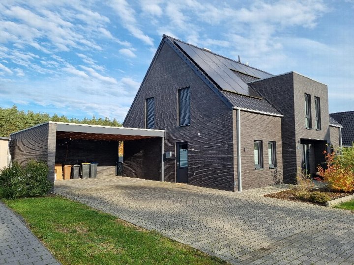 Einfamilienhaus mit dunkler Ziegelwand, Satteldach mit Solaranlage und Carport. Vorderansicht mit Eingangstür, gepflastertem Vorplatz und Grünanlagen