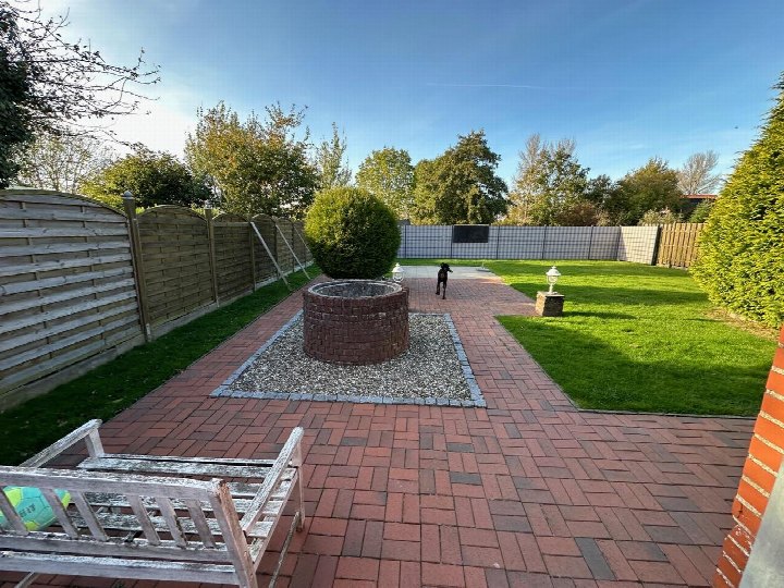 Garten mit rotem Pflasterweg, Ziegelkamin, Rasenfläche und Holzzaun. Ein Hund steht im Hintergrund