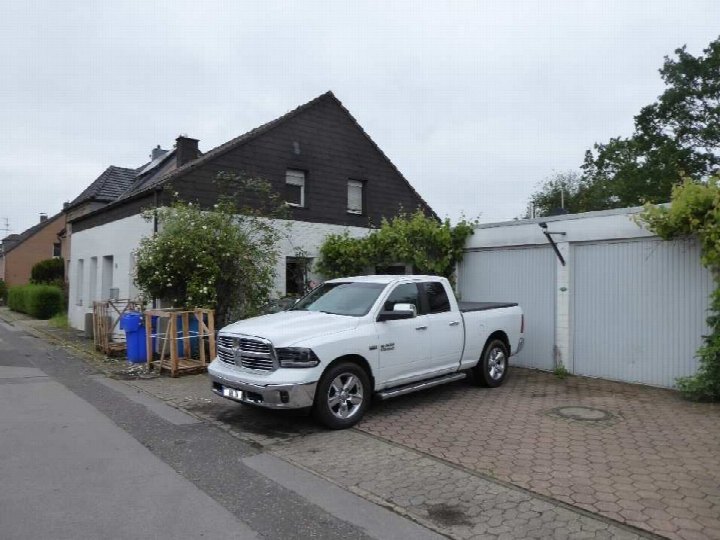 Einfamilienhaus mit dunklem Satteldach und weißer Fassade, vorne geparkter weißer Pickup, Garagenanbau rechts, gepflasterter Vorplatz