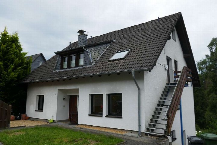 Einfamilienhaus mit weißer Fassade und dunklem Satteldach, Eingangsbereich mit Holztür, Fenster und Außentreppe zur oberen Etage, Garten im Vordergrund, Adresse Kapellenstr. 27