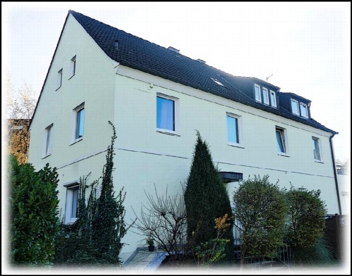 Seitenansicht eines Mehrfamilienhauses mit hellgrüner Fassade, dunklem Dach und Fenstern, umgeben von Sträuchern und Bäumen