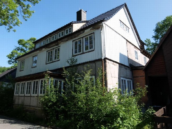 Seitenansicht eines zweistöckigen Hauses mit Holzverkleidung, mehreren Fenstern und Dachgauben, umgeben von Grünanlagen