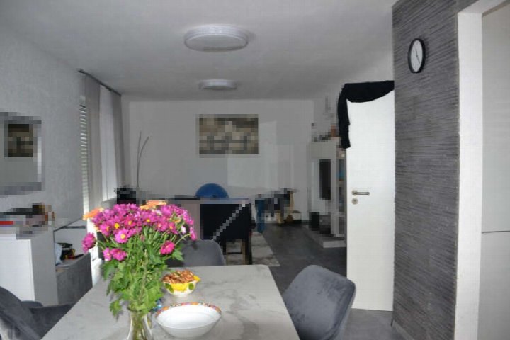 Innenraum eines Wohnbereichs mit Esstisch, Stühlen, Blumen und Blick in den Wohnraum mit TV-Regal und Wandbild