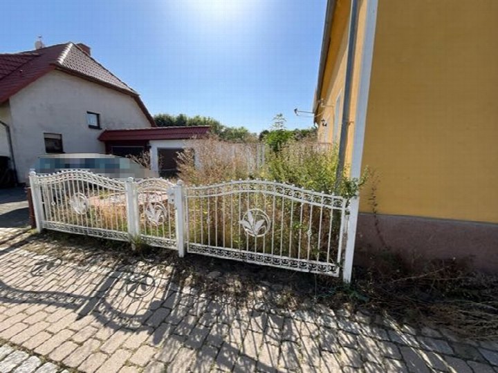 Gartenbereich mit weißem Metallzaun und überwuchertem Grün vor einem gelben Gebäude an der Weidenweg 8 in Schackensleben