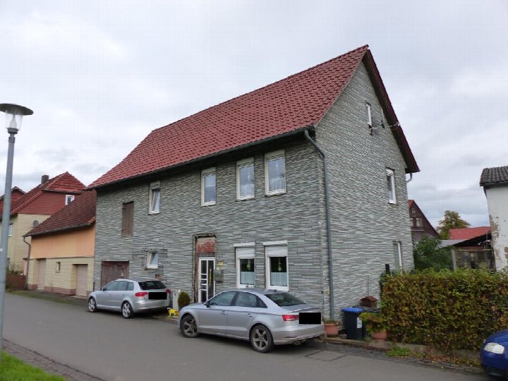 Einfamilienhaus mit grauer Holzfassade und rotem Satteldach, Eingangstür und Fenster im Erd- und Obergeschoss, geparkte Autos vor dem Haus