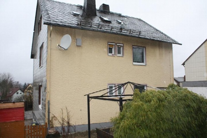 Seitenansicht eines Einfamilienhauses mit gelber Fassade, Schieferdach, Satellitenschüssel und Gartenbereich im Vordergrund