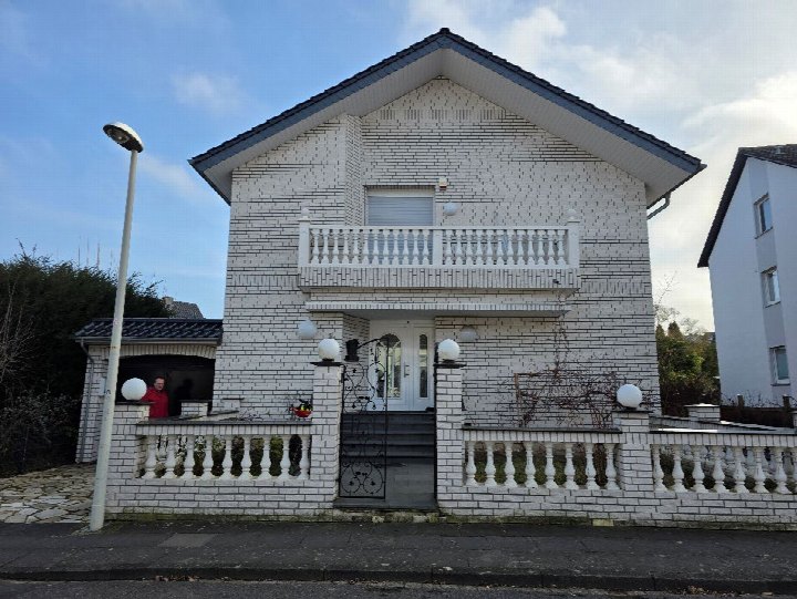 Vorderansicht eines Einfamilienhauses mit weiß verputzter Fassade, Balkon und Eingangstür. Links im Bild steht eine Person im roten Oberteil