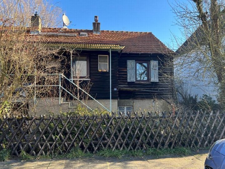 Vorderansicht eines Holzhauses mit rotem Ziegeldach, Fenstern mit Läden, Eingangstreppe und Holzzaun vorne. Lage: Bürgermeister-Beheim-Straße 3, 63165 Lämmerspiel