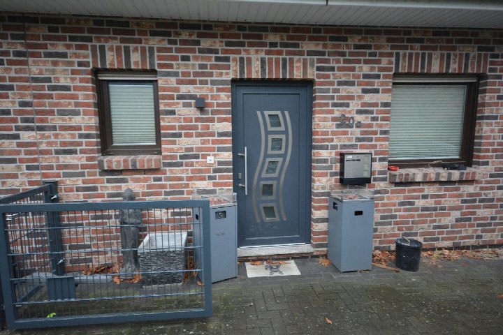 Eingangsbereich einer Doppelhaushälfte mit blauer Haustür, Backsteinfassade und zwei Fenstern mit Jalousien. Links ein Metallgitter, rechts Mülltonnen