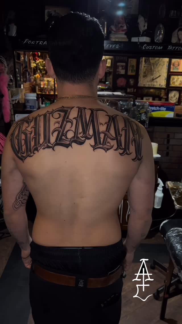 Bold Gothic-script back tattoo across the shoulders.