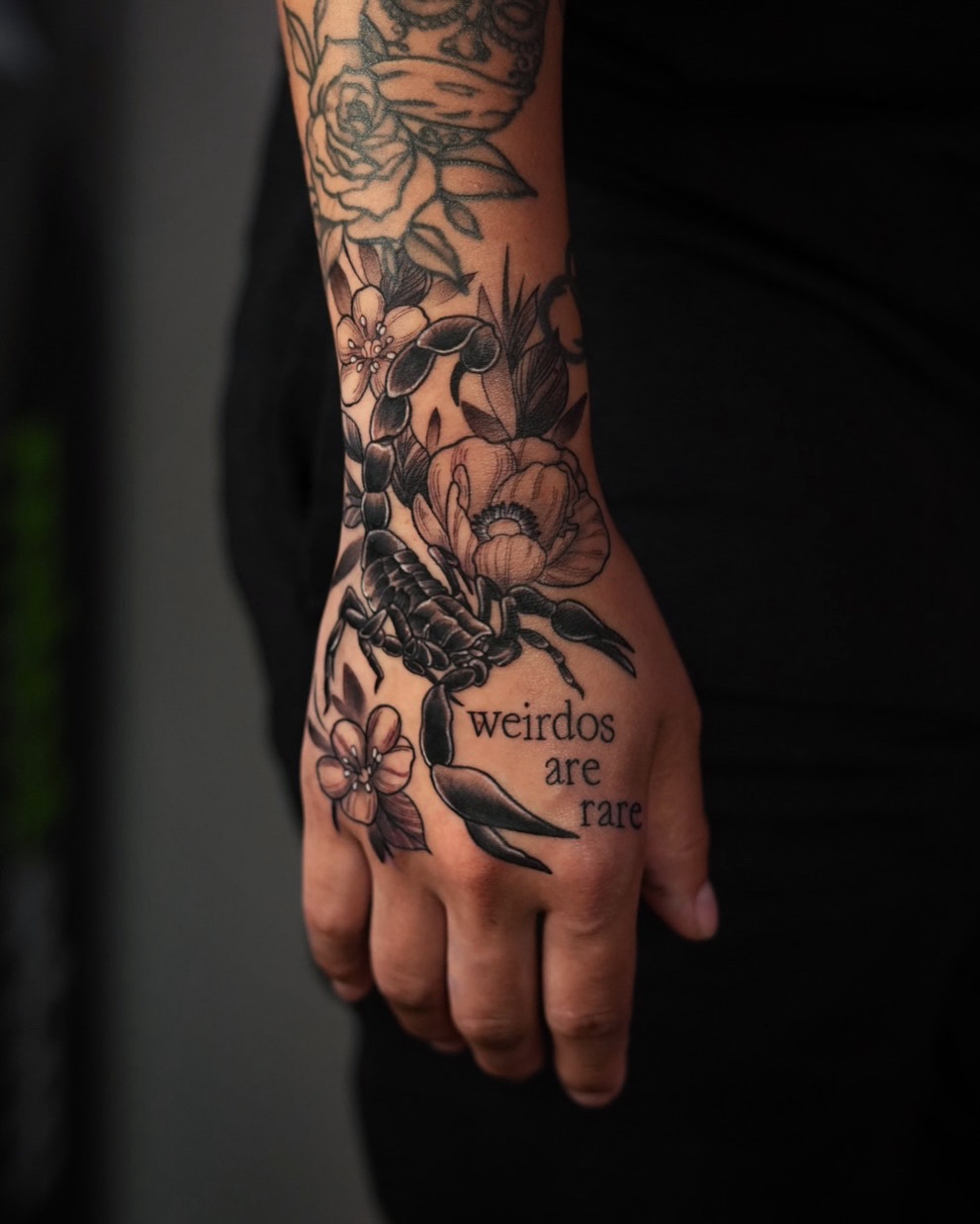 Black-and-gray botanical tattoo with a scorpion and 'weirdos are rare' text