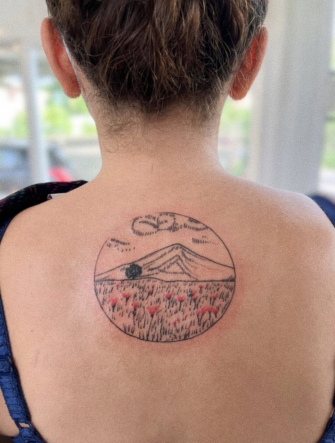 Circular landscape tattoo of mountains, field, and red flowers