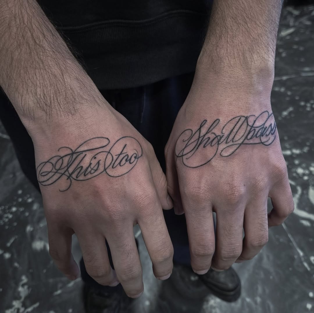 Dual-handed cursive phrase spanning wrists: 'This Too Shall Pass'.