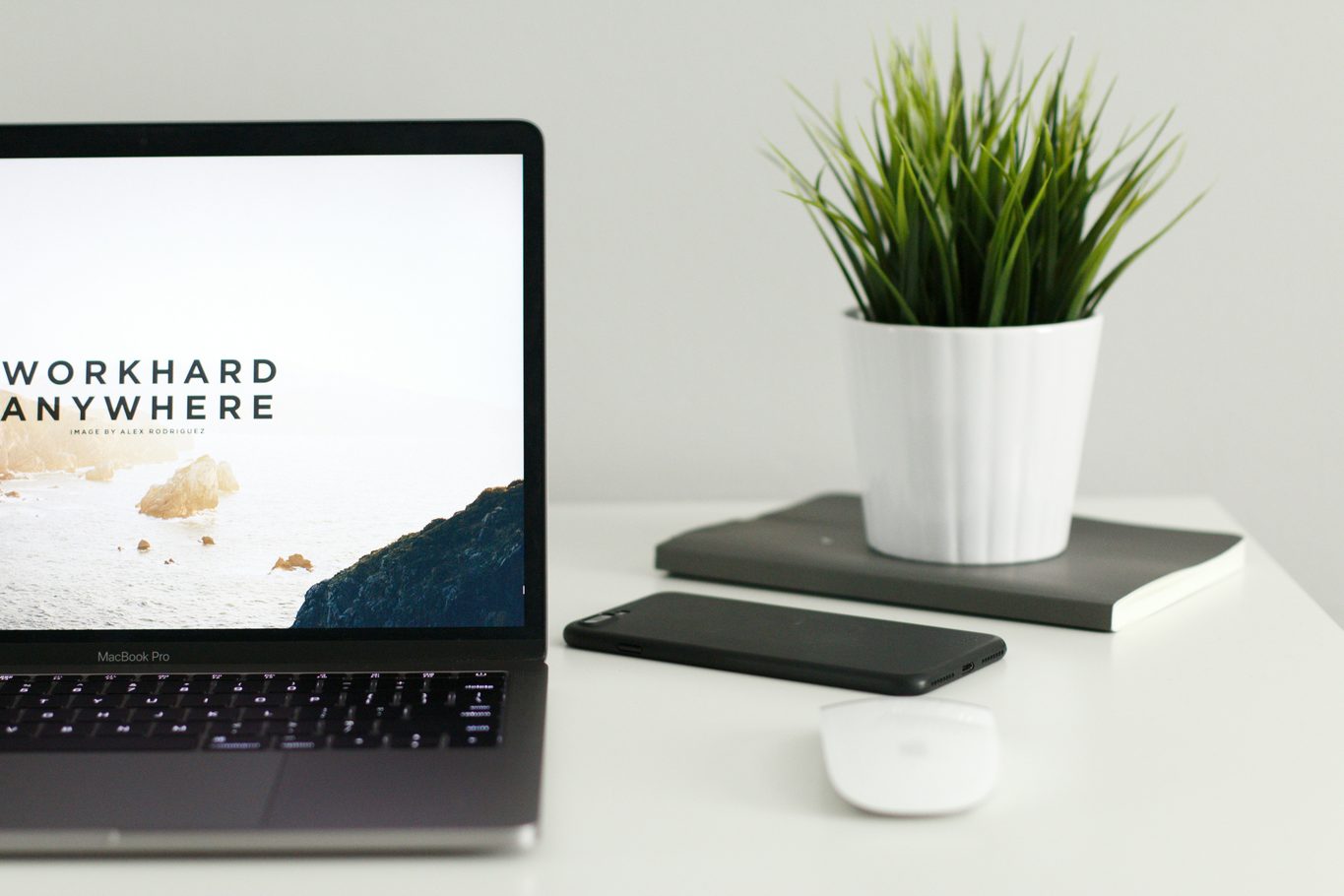 Open laptop on a white desk beside a phone, a closed notebook and a small grass plant