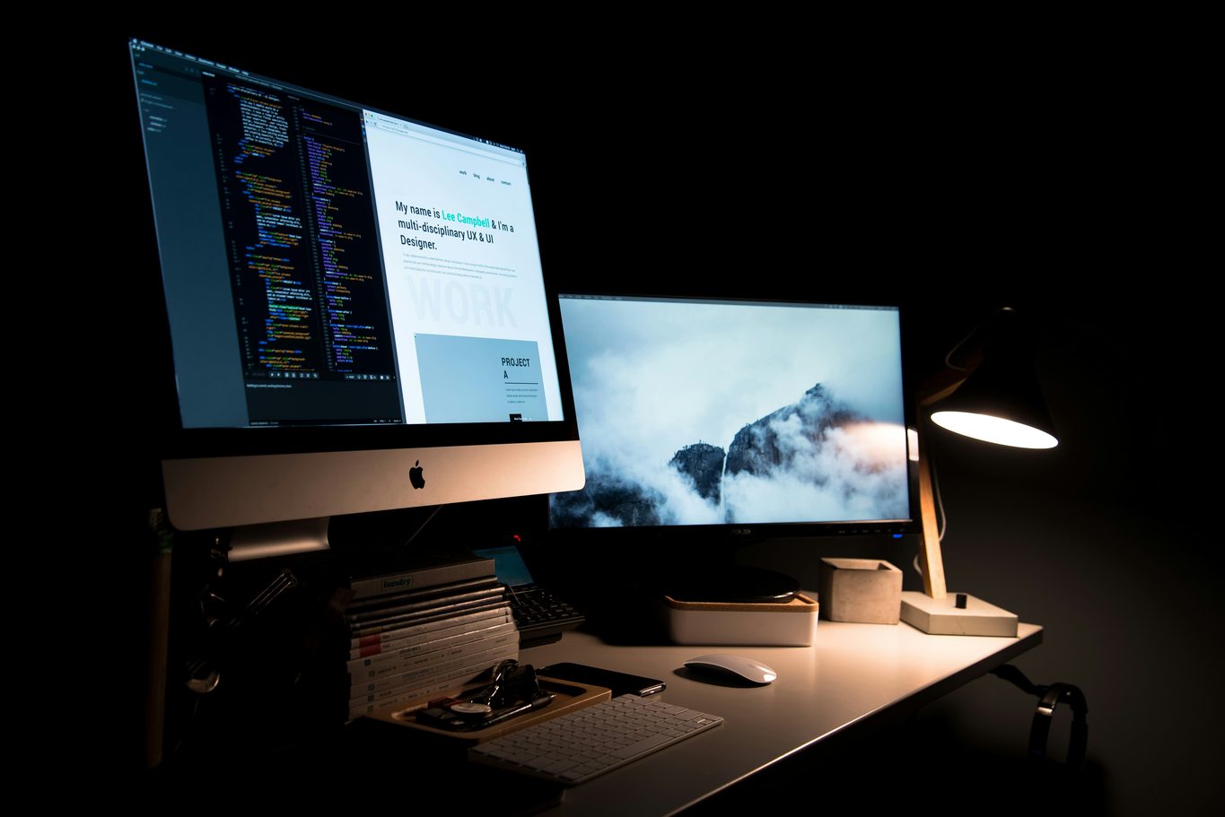 A darkened desk workstation with two lit screens and a lamp casting a narrow pool of light