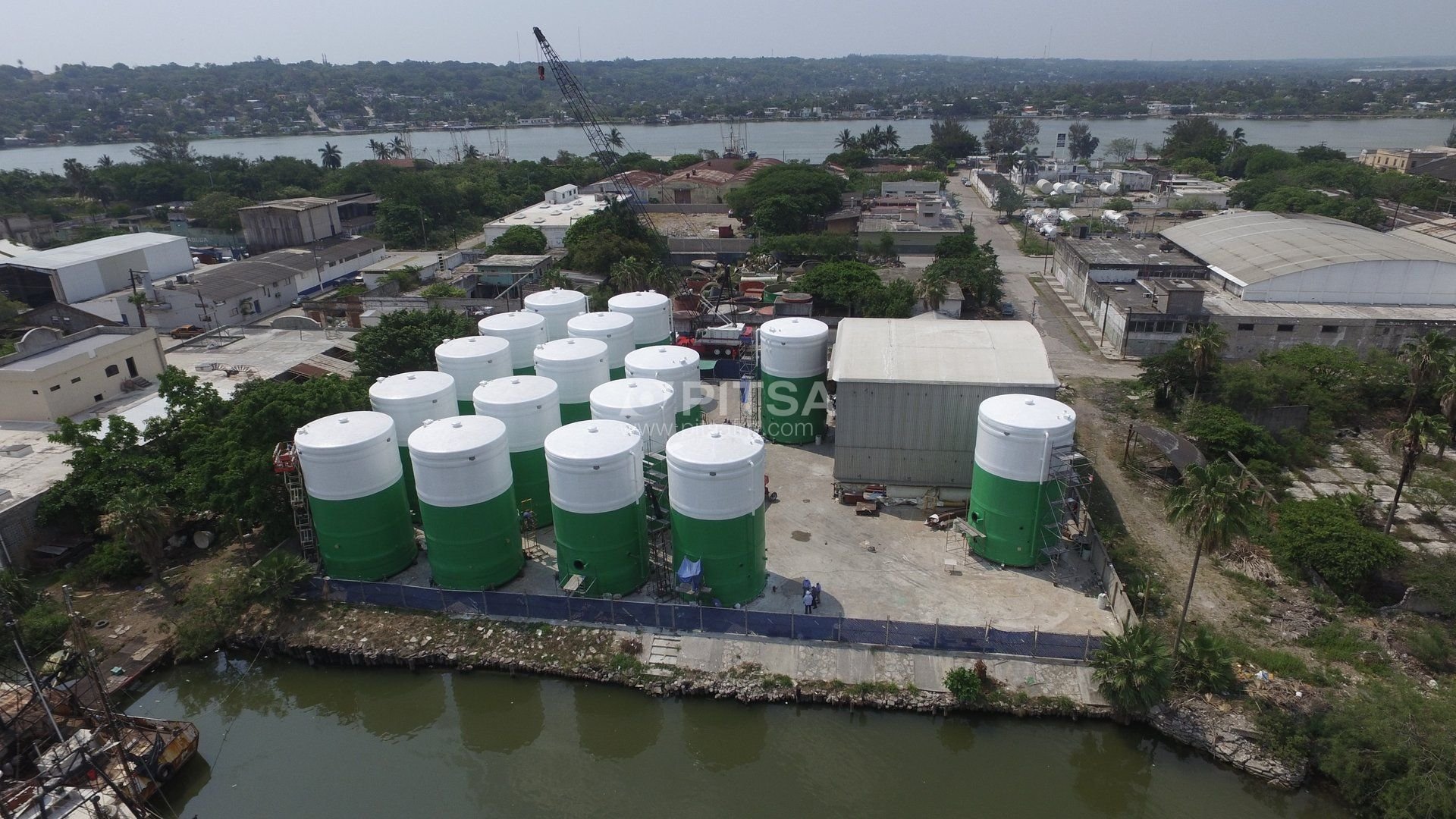 Aerial view of twelve large green and white FRP storage tanks at an industrial facility near a waterfront, surrounded by trees and buildings.